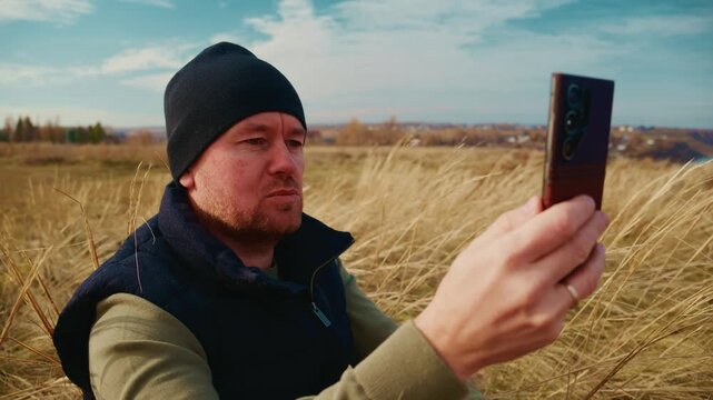 A thoughtful man sitting in a grassy field holds his smartphone, gazing intently at the screen as he enjoys a serene moment in nature, surrounded by tall grass with a distant landscape under a blue