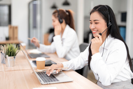 Smiling Asian woman wearing a headset with a microphone, providing excellent customer support or attending a virtual meeting at her laptop in a modern call center or office.