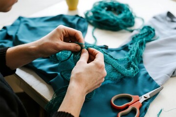 Hands knitting teal yarn onto a garment with scissors and fabric nearby