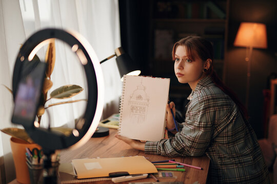 Portrait of Caucasian teenage girl sitting at desk holding sketchbook with architectural drawing, looking into camera, surrounded by art supplies and using smartphone with ring light
