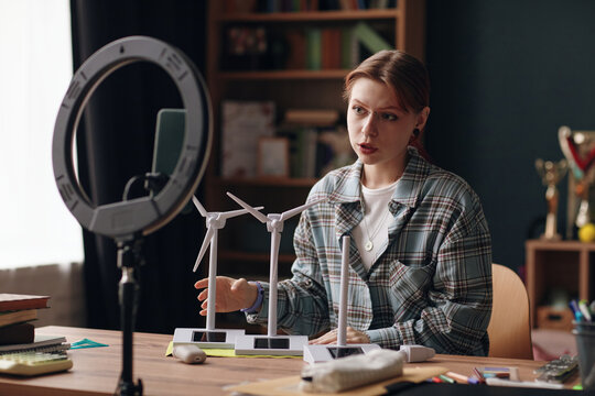 Caucasian teenage girl explaining wind turbine models while recording educational video with smartphone and ring light in bedroom, demonstrating interest in science and technology projects