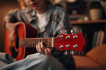 Caucasian teenage girl sitting in room playing acoustic guitar, focusing on hand strumming strings, relaxed posture, casual clothing, blurred background with shelves and plants