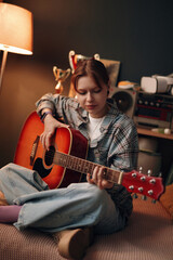 Obraz premium Caucasian teenage girl sitting on bed playing acoustic guitar in bedroom, focusing on strumming strings, surrounded by personal items and trophies, engaging in musical activity