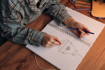Caucasian teenager drawing architectural sketch in notebook using colored pencil, sitting at wooden desk with colored pencils and digital tablet nearby, focusing on creative activity