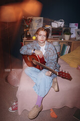 Portrait of Caucasian teenager girl sitting on bed holding acoustic guitar, looking into camera,...