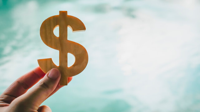 Hand holding wooden dollar sign over blurry water background, symbolizing financial risk, money instability, uncertain investment, cash liquidity, and economic volatility in personal finance. - Powered by Adobe