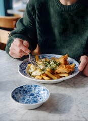  a man in a green sweater eating from a white porcelain plate with fish and chips, his hands holding a fork