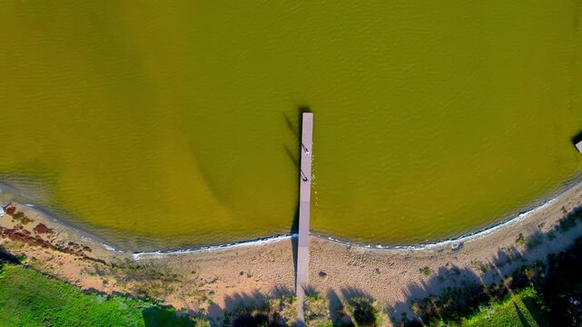 High top-down aerial shot of the jetty at Siloam Pool, showing the yellow water, a couple running along the jetty, and their long shadows on the surface. A unique and visually striking coastal scene.