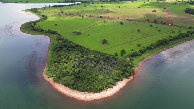Vibrant aerial shot of a forested peninsula jutting into a massive reservoir in Mato Grosso do Sul, Brazil. Lush green farmland stretches across the land under bright, sunny conditions.