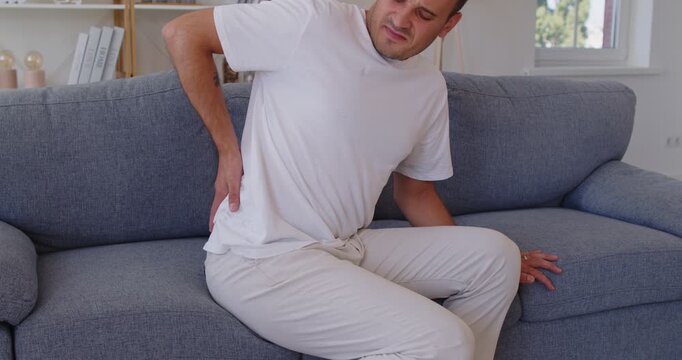 Sick man with pain in spine or lumbar sitting on sofa. Sad person in white tee shirt holding lower back, leaning to stand from couch. Patient with face tense suffering from backache and cramps