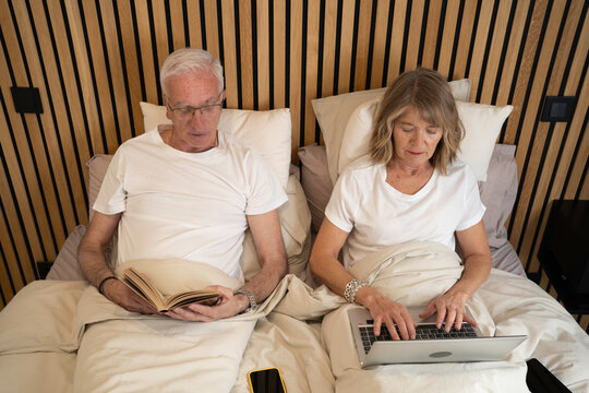 Top view of senior couple enjoying leisure time in bed, one reading a book, the other using a laptop