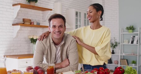 Happy diverse couple cooking breakfast together in bright kitchen. Man and woman preparing healthy food with bread, juice, salad and fresh fruits on countertop. Home daily routine and relationships - Powered by Adobe
