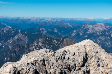 The rugged limestone summit of Jof di Montasio overlooks endless layers of blue mountain ridges, offering a breathtaking panorama of the Carnic Alps and Dolomites under a clear sky in Italy.