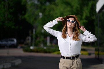 Elegant business woman in sunglasses enjoys sunny day on city streets. Chic city traveler, sophisticated business woman, holds onto her hat, looks at sun, and smiles.