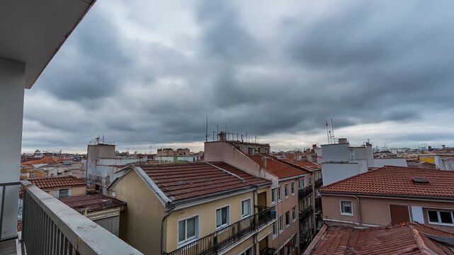 City view from a balcony showing a densely packed neighborhood with terracotta roofs under a dramatic cloudy sky, capturing a theme of urban landscape in a time lapse sequence.