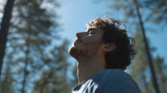 A young man stands among tall trees gazing upwards with his eyes closed embracing the warmth of sunlight. He appears to be meditating or reflecting peacefully surrounded by nature.