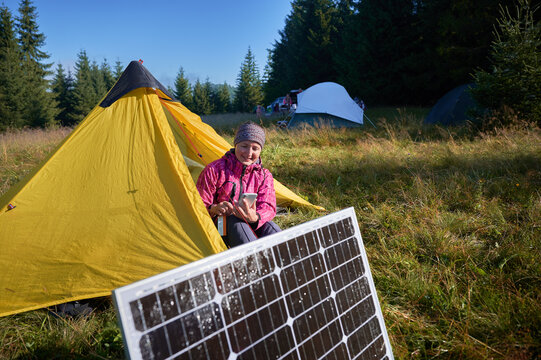 Happy woman sits in front of yellow tent, charging her phone by using solar panel, highlighting use of renewable energy and eco-friendly camping. On background lush green trees and clear blue sky.