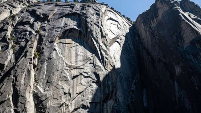 Jagged cliffs of grey stone rise steeply under a clear blue sky shadowed by dramatic vertical features