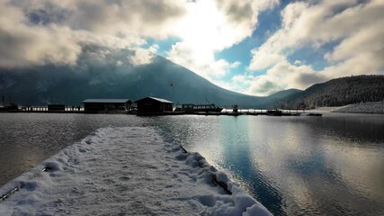 Clear mountain lake stretches from a snow edged dock toward rugged snowy peaks wrapped in drifting clouds, under bright blue sky and soft winter light in Alberta, Canada. - Powered by Adobe