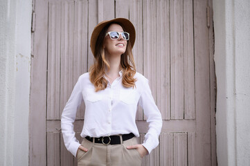 Fashionable traveler in hat and sunglasses stands in front of wooden doors in Europe. Stylish woman on historic city street poses in front of weathered door for lifestyle photo.