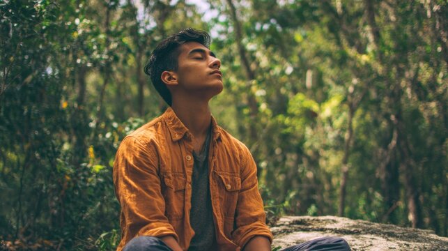 A young man sits cross legged on a rock eyes closed and face relaxed while meditating in a lush green forest filled with trees and natural light.
