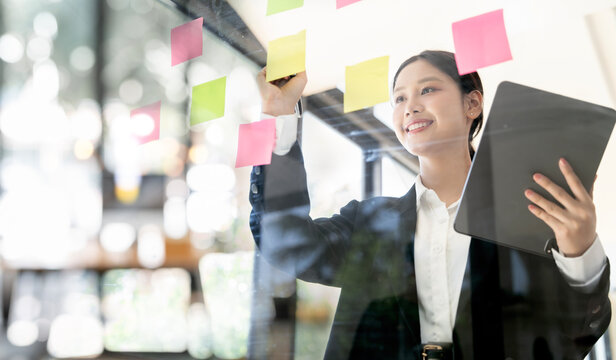 Smiling Asian businesswoman holding a tablet and attaching sticky notes to a glass wall, demonstrating organization, planning, and brainstorming in a modern office. - Powered by Adobe
