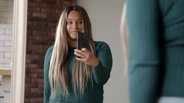 Portrait of cheerful cubby black woman in stylish clothes posing in front of mirror, taking photo on mobile phone for social media while spending leisure in domestic room.