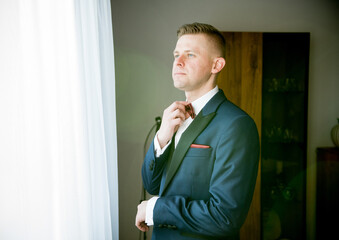 Elegant groom in navy blue suit adjusting bow tie while standing by bright window indoors