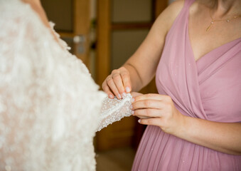 Bridesmaid in pink dress fastening delicate beaded lace sleeve on bride’s wedding gown indoors