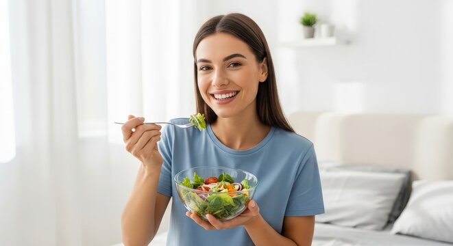 Smiling young woman eats fresh vegetable salad with fork in bright home on a sunny day
