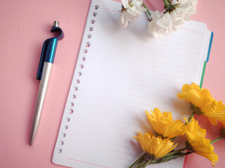 Notebook and pen. Flat lay of blank spiral notebook, blue pen, and yellow chrysanthemum flowers on pink background with copy space. Workspace mockup with open diary, stationery, and floral arrangement