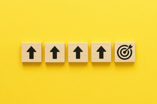 Wooden blocks with upward arrows and a target icon against a yellow background, symbolizing growth, achievement, and goal-oriented mindset in business and personal development.