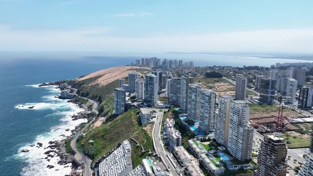 Backward drone aerial of Vi&ntilde;a del Mar clifftop towers along rocky Pacific coastline with sand dunes and turquoise ocean waves, Chile resort