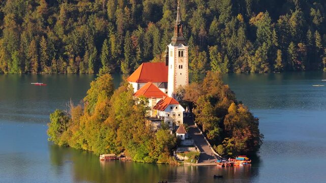 Aerial view of lake Bled with of Cerkev Marijinega Vnebovzetja with colored trees in forest. Rowers on boats.mov