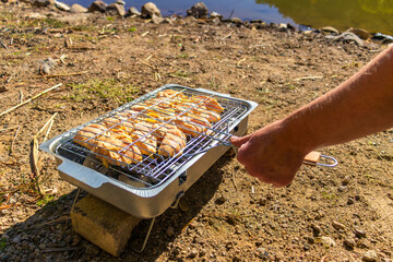 Chicken cooks on a portable grill by a lake during a hiking and outdoor travel stop. 