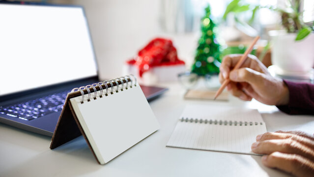 close up of calendar, notebook with computer laptop, red hat, christmas tree for decoration on desk and blurred hand holding pencil taking note on background, christmas holidays concept - Powered by Adobe