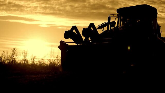 Simple rural life and works in countryside, farmer driving bulldozer in farmland . Backlit silhouette of worker riding contemporary tractor in nature, beautiful sunset or sunrise, modern equipment