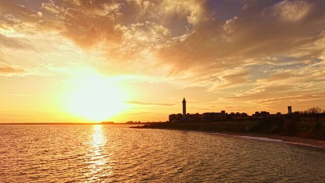 As the sun sets on the horizon, warm golden hues reflect off the tranquil ocean while a lighthouse stands tall in the background, framing a peaceful shore.