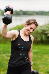 Fototapeta premium Smiling woman lifting dumbbell by lake in grassy park