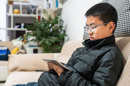A young boy is sitting on a sofa using a digital tablet, engaged in an activity indoors.