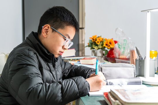 A young boy wearing glasses is doing homework carefully at a desk with various stationery items around. - Powered by Adobe