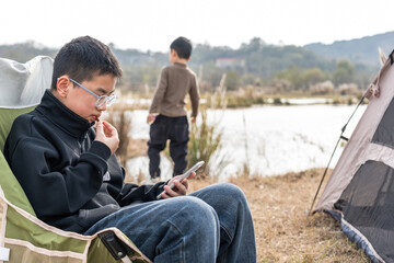 A boy sits on a camping chair using a mobile phone by a tent near a body of water in an outdoor setting.