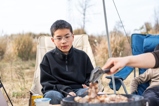A young boy is having a good time camping outdoors, with someone cooking food for him.