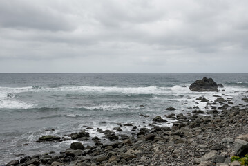Waves rolling onto a rocky shore in Tenerife, with dramatic cliffs and cloudy skies in the background
