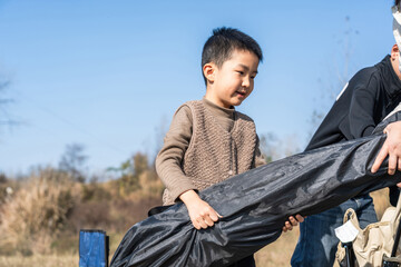 A young boy is seen assisting in preparing an outdoor activity, holding a black bag in a natural outdoor setting.