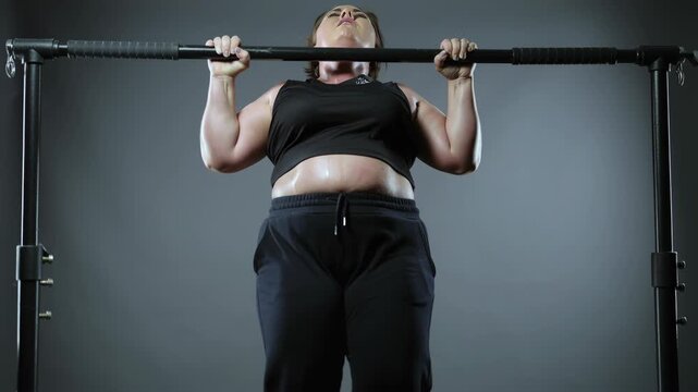 Woman pull up on horizontal bar. Overweight lady performing physical exercise. Strength training and hard workout concept.