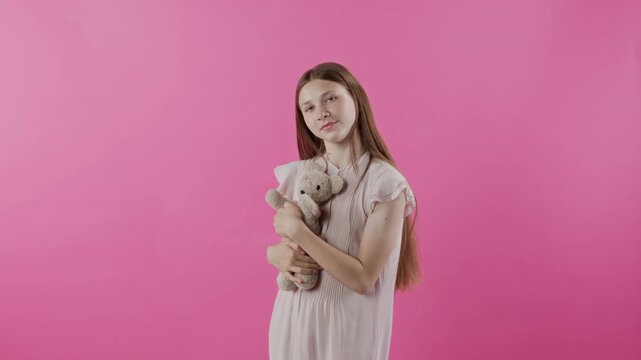 Studio portrait of playful teenage girl with long hair holding teddy bear up then turning to camera with joyful smile while posing on pink backdrop, concept of childhood