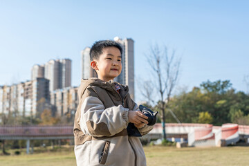 Fototapeta premium A young boy is holding a camera while standing outdoors in an urban area with buildings in the background.