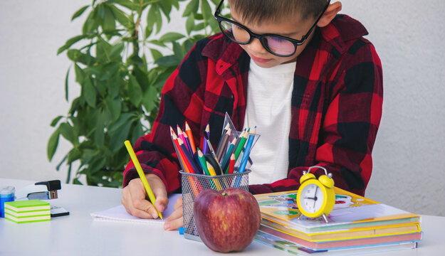 Child writing or drawing at a desk surrounded by school supplies, books, an apple,and an alarm clock - Powered by Adobe