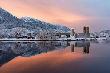 A factory stands by a tranquil lake with snow-covered mountains in the background. The sky is lit with warm colors, reflecting beautifully on the water surface during dusk.